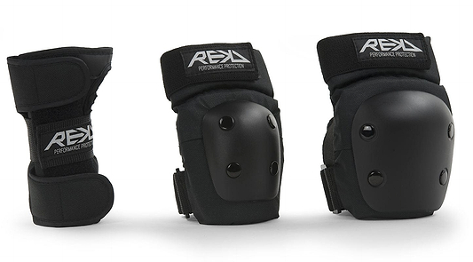 Three black protective gear items from REKD are showcased: an ergonomic wrist guard on the left and two knee or elbow pads on the right. The REKD Junior Heavy Duty Triple Skate Protection Pad Set includes hard plastic shells with the "REKD" logo on the straps, providing protection for extreme sports enthusiasts.