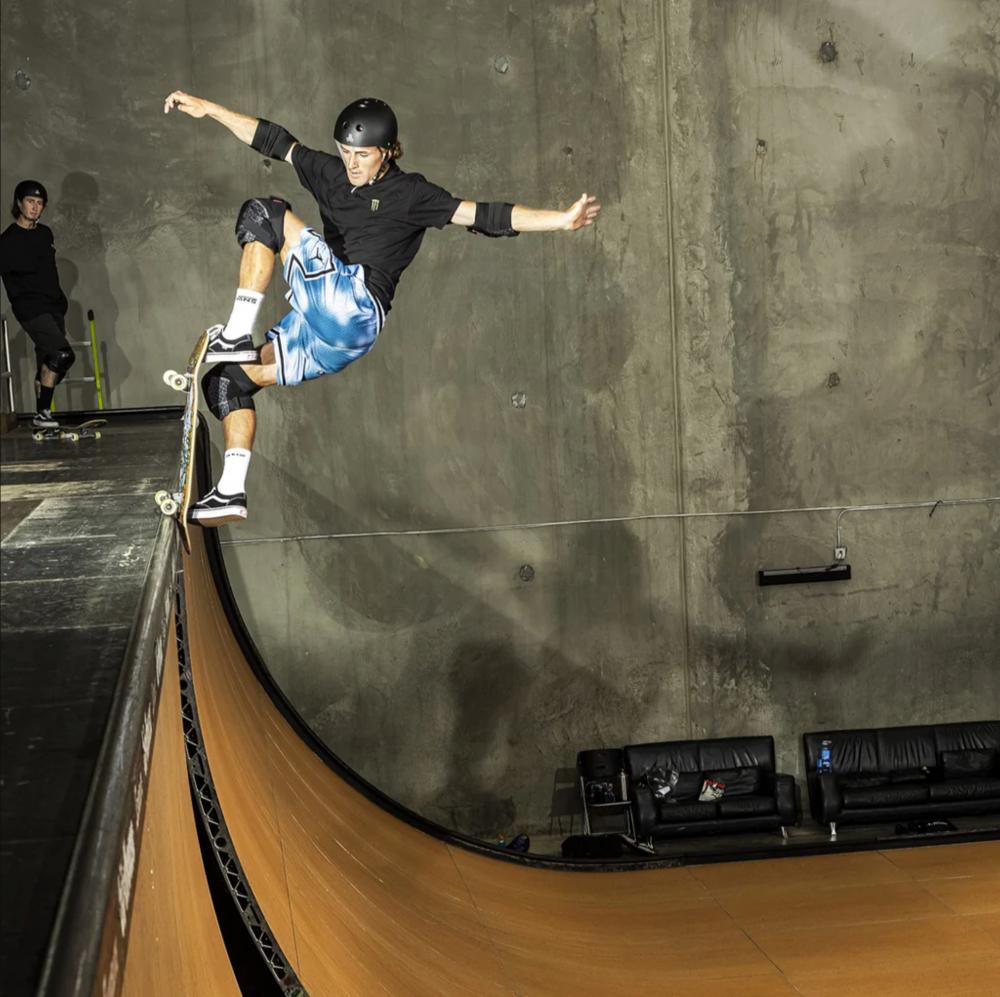 A skateboarder wearing a helmet, black shirt, blue shorts, and the 187 x Independent Killer Adult Combo Knee & Elbow Pad Set performs a trick on an indoor vertical ramp while another person in protective gear stands in the background.