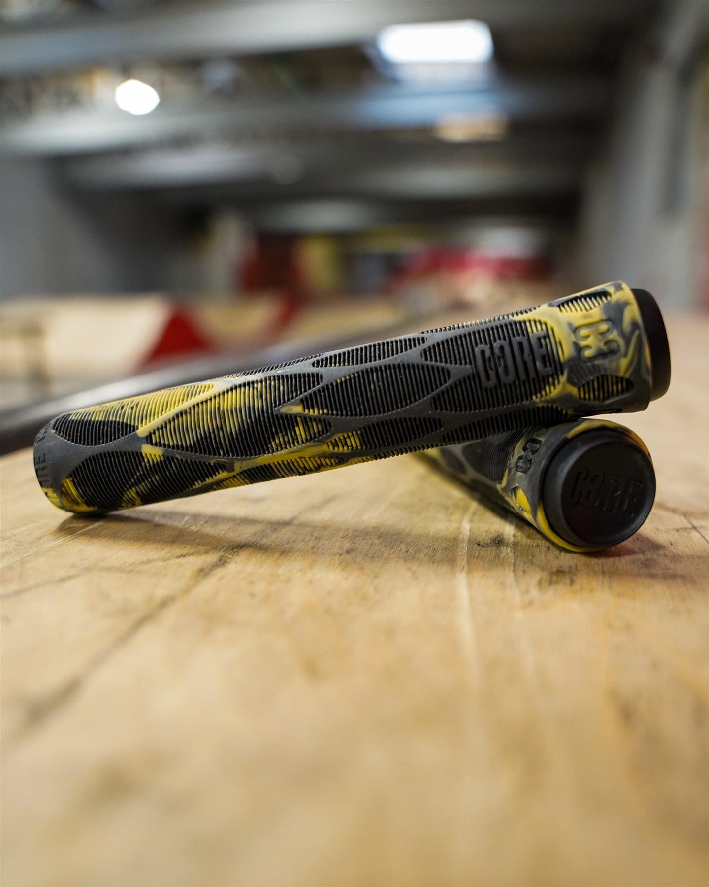 Close-up of CORE Pro Bark Black / Gum Stunt Scooter Grips (170mm) by CORE, featuring shock-absorbing design and visible bar ends, displayed on a wooden indoor surface with a softly blurred background.