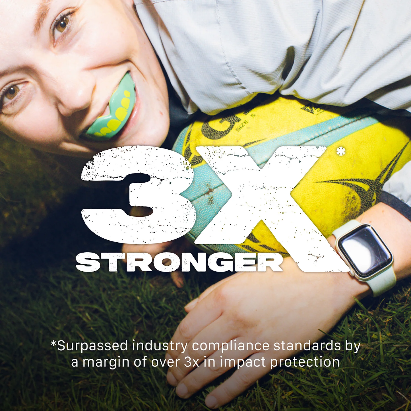 A smiling person with a bright green Safejawz Extro Series Protection Mouthguard - Ogre lies on grass, holding a yellow ball. Bold text says "3X STRONGER." Small print notes it exceeds industry compliance by over 3x in impact protection.