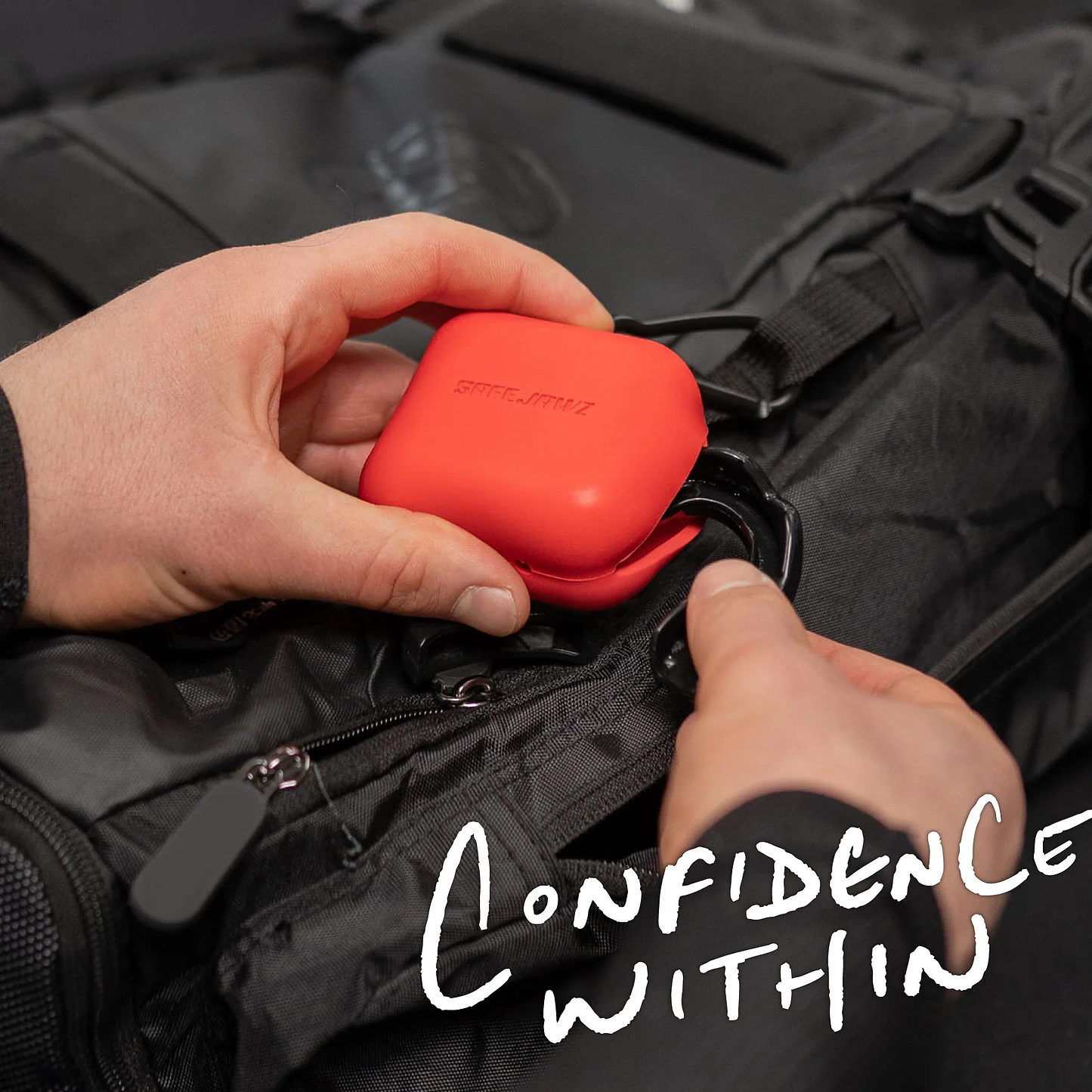 A person clips a red Safejawz Premium Silicone Mouthguard Case onto a black backpack, featuring the Safejawz logo and "CONFIDENCE WITHIN" in white near the bottom right corner.