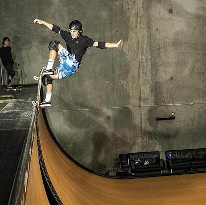 A skateboarder wearing a helmet, black shirt, blue shorts, and the 187 x Independent Killer Adult Combo Knee & Elbow Pad Set performs a trick on an indoor vertical ramp while another person in protective gear stands in the background.