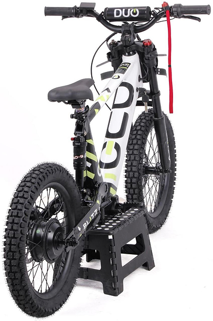 The Duo 18" Blitz Kids Electric Balance Bike in white and black, by Duo, sits on a black step stool. It features chunky tires, a padded seat, and a red strap on the handlebars, shown from the rear angle.