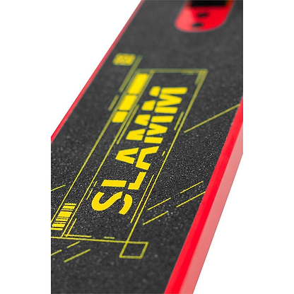 A close-up of the Slamm Classic Mini Complete Stunt Scooter reveals its skateboard-like deck covered with black grip tape featuring the brand name "SLAMM" printed in vibrant yellow, capturing the bold energy typical of freestyle scootering. The edges boast a striking red color, with glimpses of an intricate graphic design visible through the grip tape.