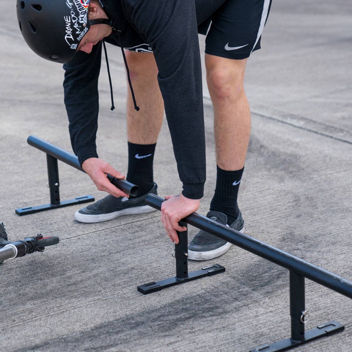 A helmeted person in a black hoodie, shorts, and sneakers adjusts a Madd Gear MGP Ninety9 Grind Rail (251cm) on concrete, likely prepping for grind practice while skateboarding or biking.