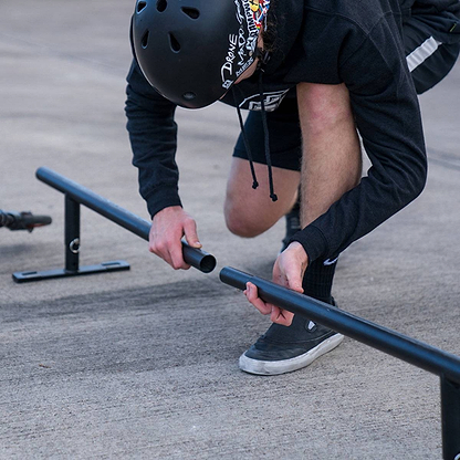 A person in a helmet and hoodie kneels to assemble the Madd Gear MGP Ninety9 Grind Rail - 251cm, getting ready for practice grinds.