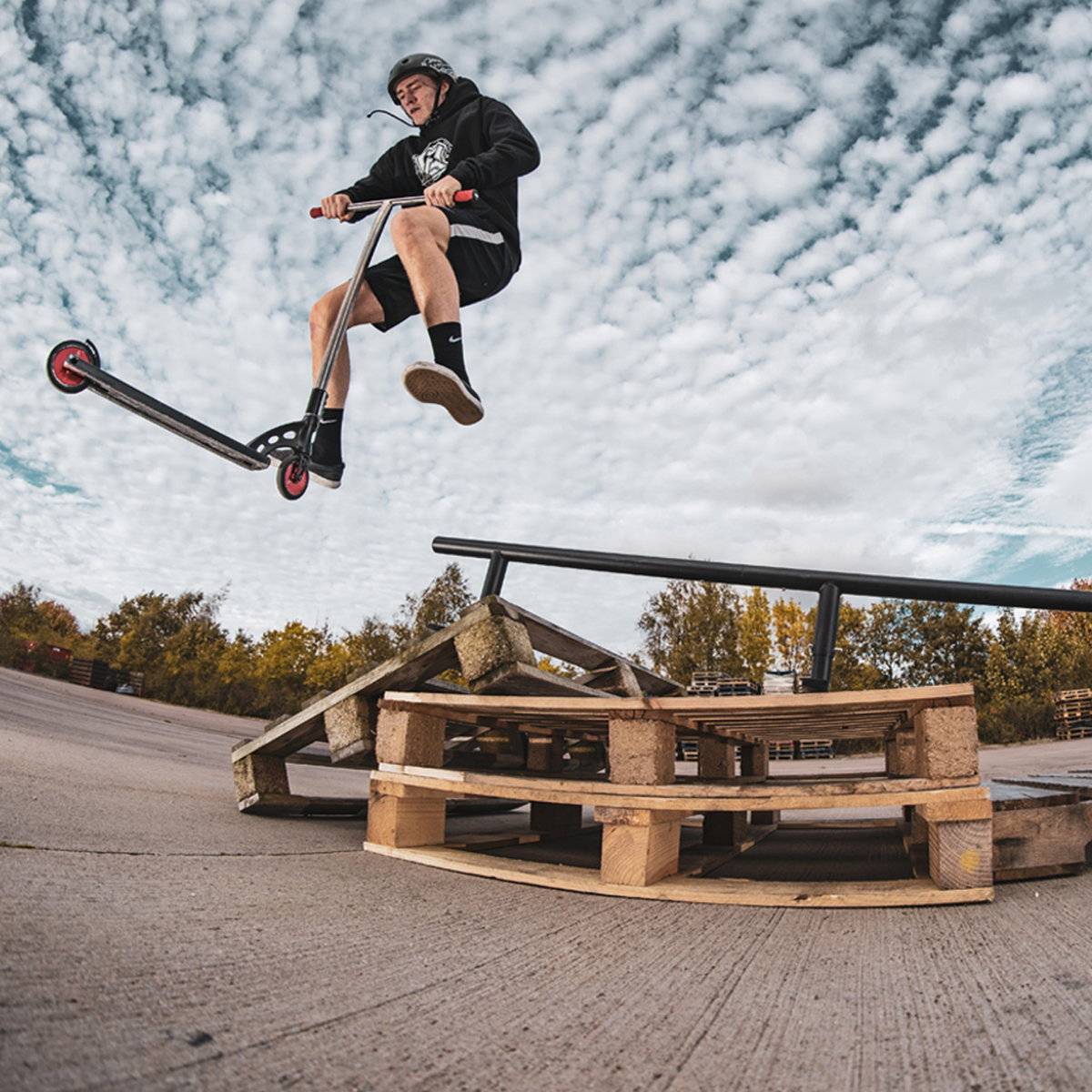 A person wearing a helmet jumps on a scooter over stacked pallets at an outdoor skate park, practicing grinds on the Madd Gear MGP Ninety9 Grind Rail - 251cm, with cloudy skies and trees in the background.
