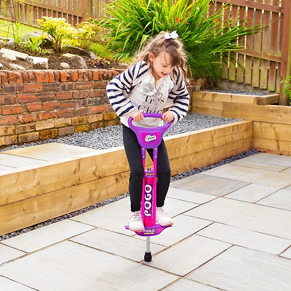 A smiling young girl with long hair, wearing a striped jacket and black pants, plays on an Ozbozz Kids 89cm Pogo Stick - Pink/Purple in a paved backyard with raised garden beds and a wooden fence.