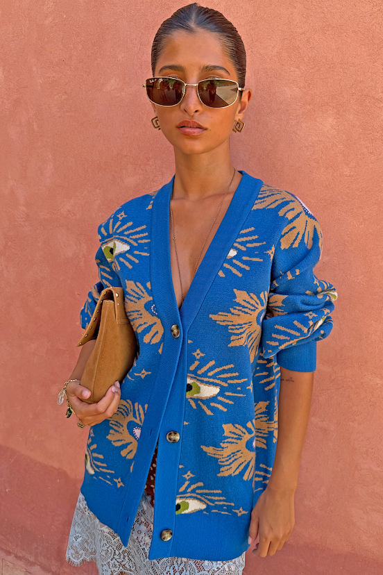 Model wearing the relaxed Blue Eye Cardigan with an eye motif and heart illustrations against a pink wall.