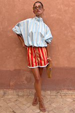Blue Stripe Batwing Blouse with high neck and wide sleeves, styled with red shorts against a terracotta wall.