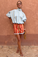 Blue Stripe Batwing Blouse with high neck and wide sleeves, styled with red shorts against a terracotta wall.
