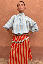 Blue Stripe Batwing Blouse with voluminous sleeves and high neck, worn by a model in front of a terracotta wall.