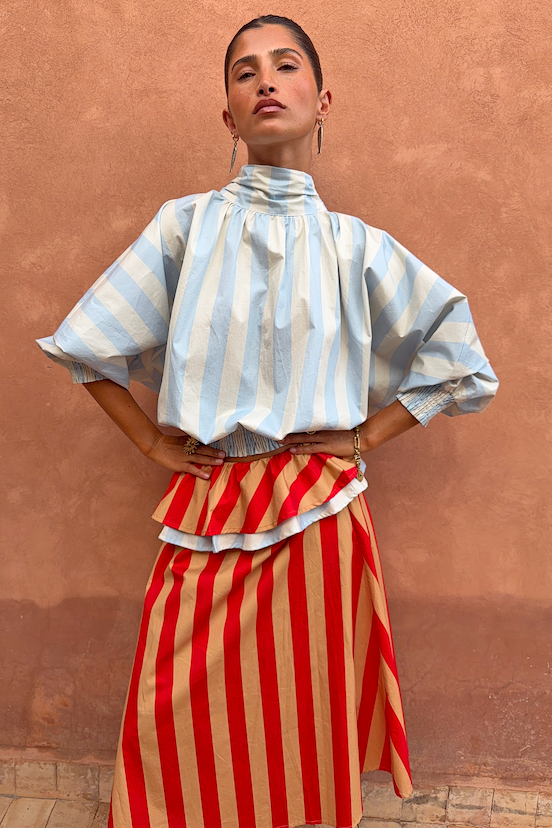 Blue Stripe Batwing Blouse with voluminous sleeves and high neck, worn by a model in front of a terracotta wall.