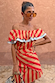 Red, tan and light blue Tomato Stripe Scarf styled as a multi-wear ruffled top against a terracotta wall.