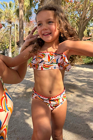 Bambina che indossa il bikini Banana multicolore con top a volant e bordini rossi in un set tropicale.