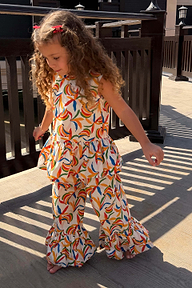 Bambina con coordinato in cotone crema a stampa banane, top peplo e pantaloni svasati su un patio soleggiato.