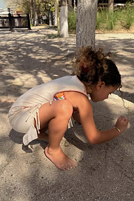 Bambina con abito in jersey di cotone panna e stampa a banane con frange, che gioca nella sabbia.