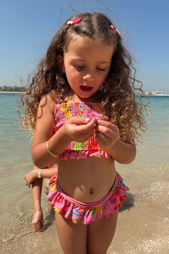 Young girl wearing the Girls Pink Fruit Crop Bikini Set with a shirred bandeau top and ruffle bottoms at the beach.