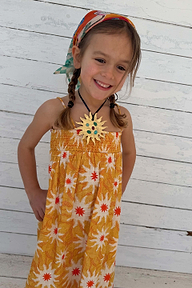 Girl wearing a yellow cotton maxi sun dress with a sun print and shirred bodice against a wooden wall.