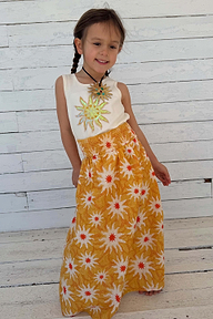 Child model in a yellow sun print cotton maxi dress with a shirred waist against a white wooden wall.
