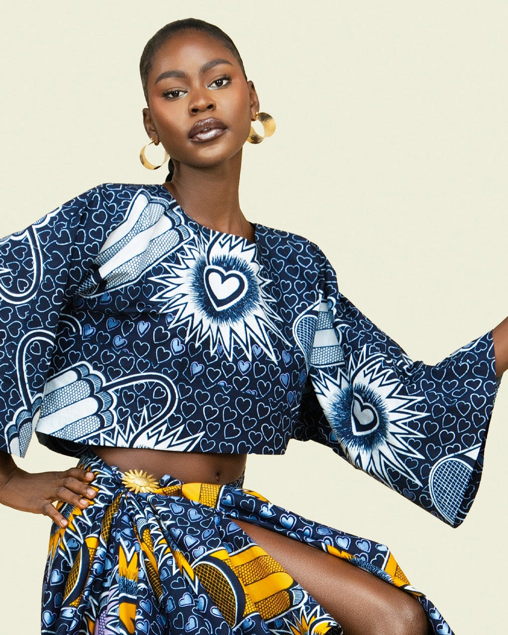 A model wearing a matching two-piece African wax print set with a navy blue and yellow geometric pattern, set against a solid light beige background.