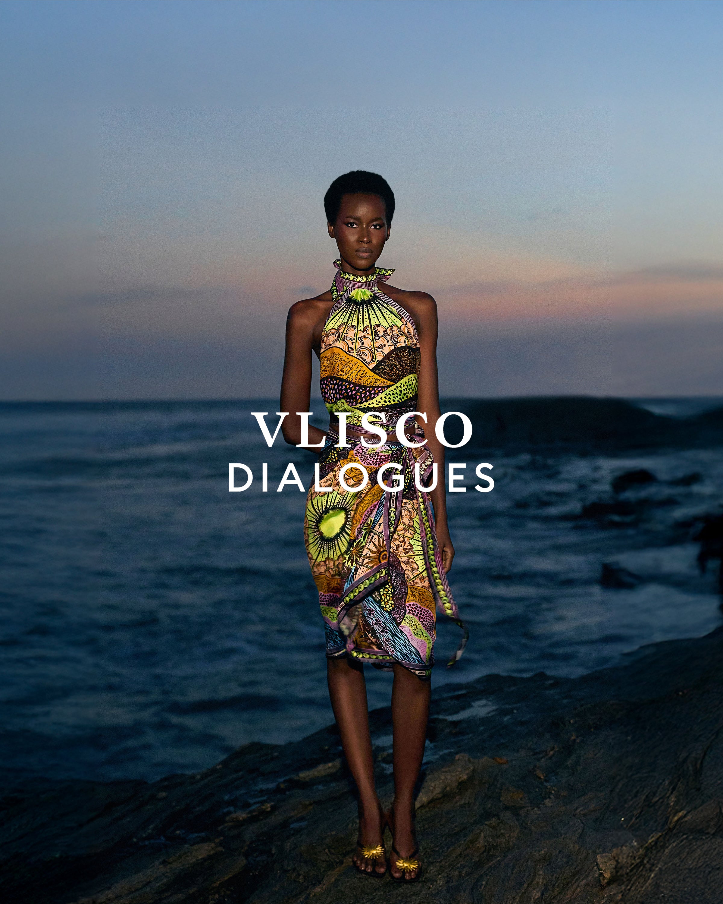 Vlisco Dialogues model standing on rocks by the sea at dusk wearing a patterned dress