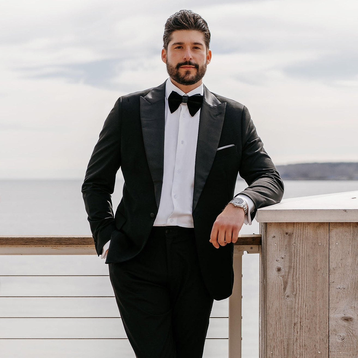 Man in a black tuxedo standing on a wooden deck with water and sky in the background