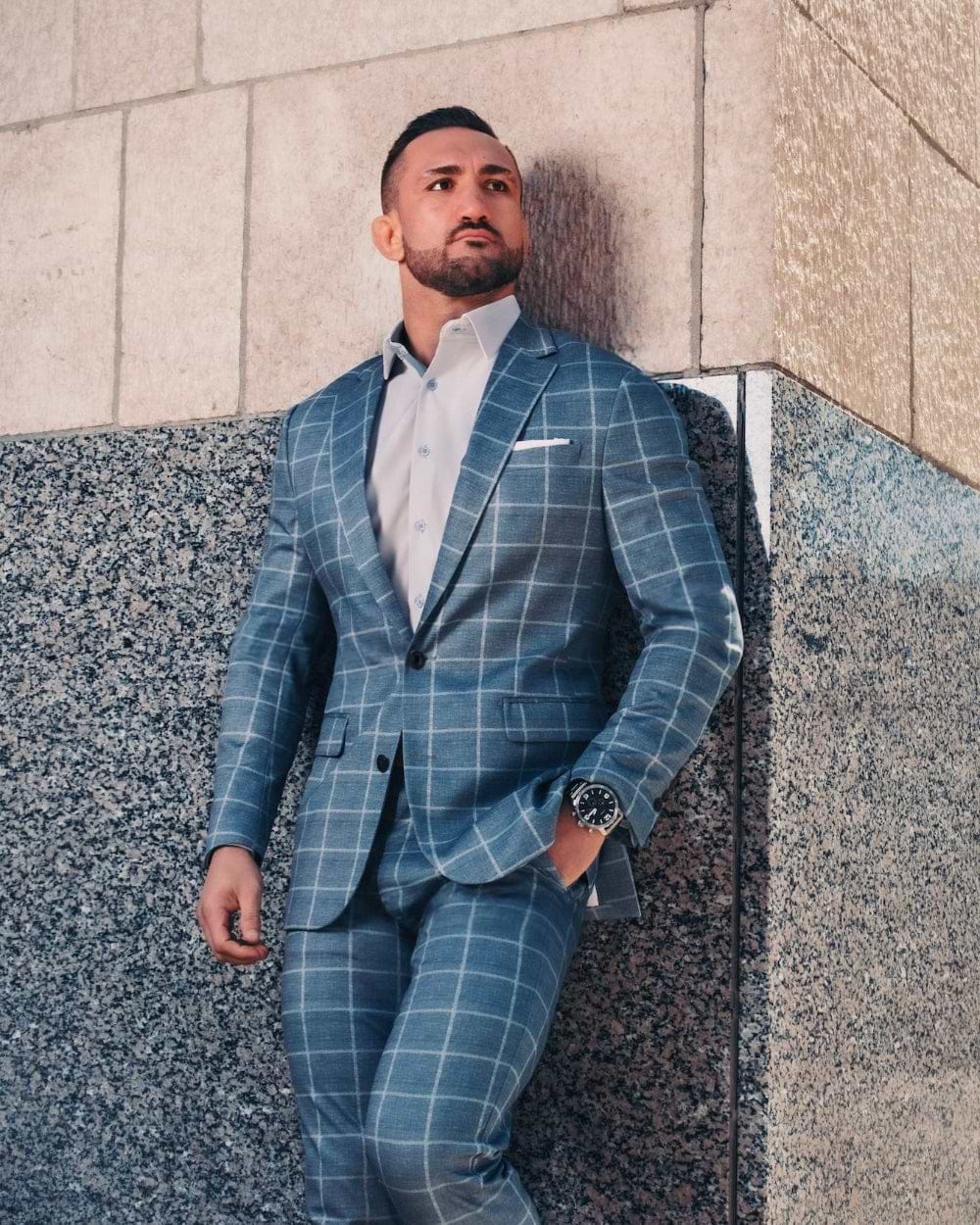 Man in a blue checkered suit standing against a building wall.
