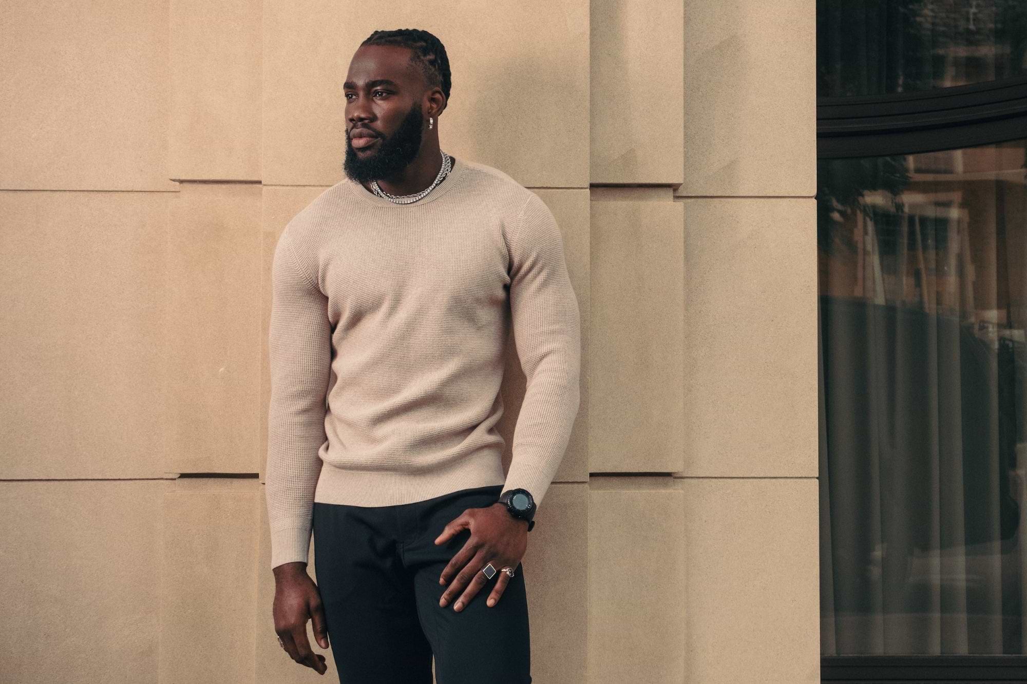 Man wearing a beige sweater and black pants standing against a modern building.