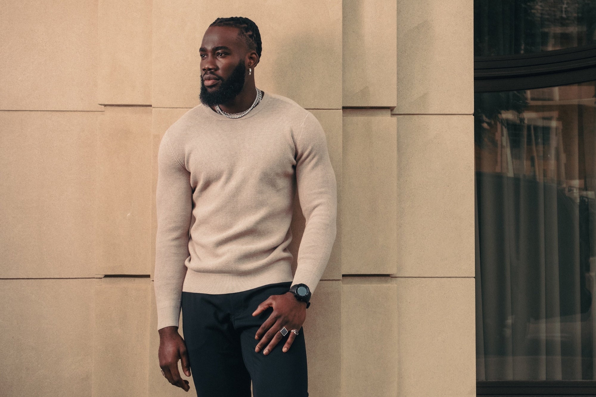 Man wearing a beige sweater and black pants standing against a modern building.