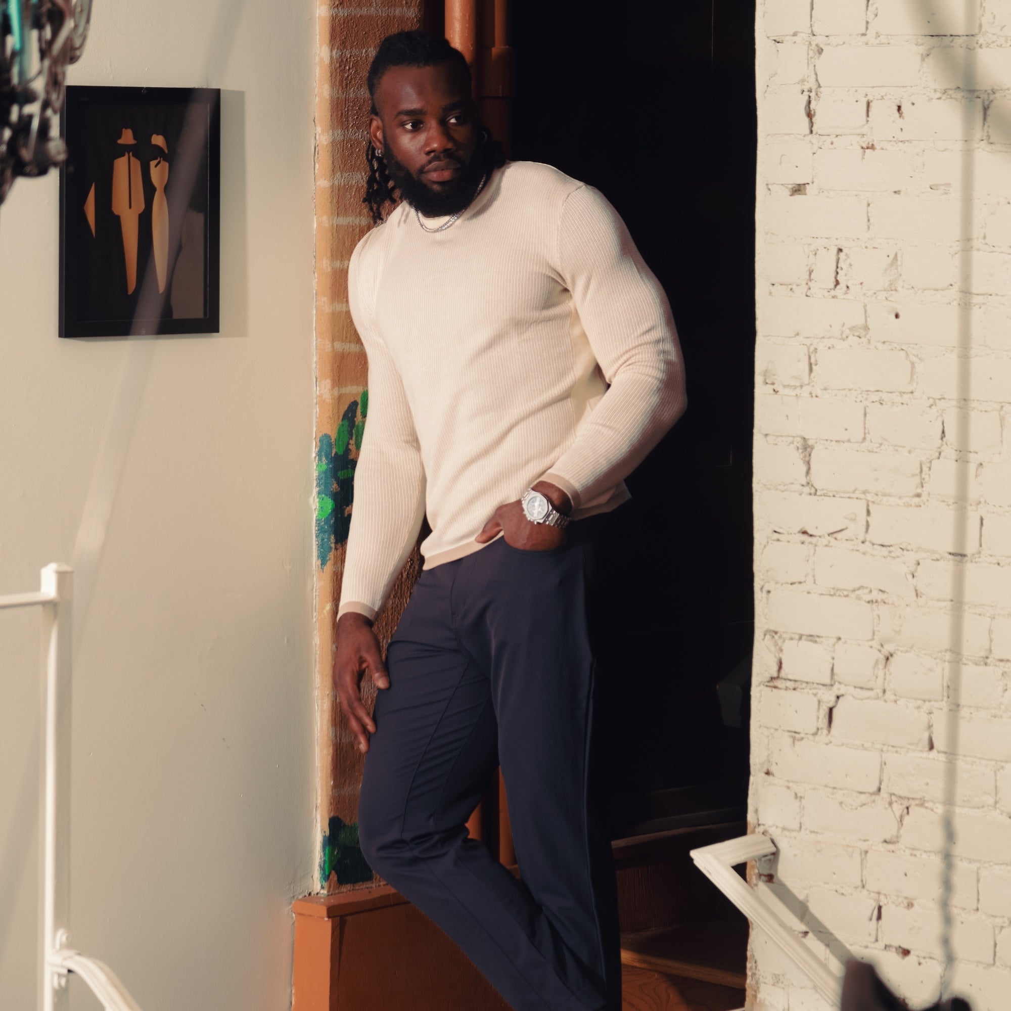 Man in a white sweater and navy pants standing on a staircase with a white wall and brick wall in the background.