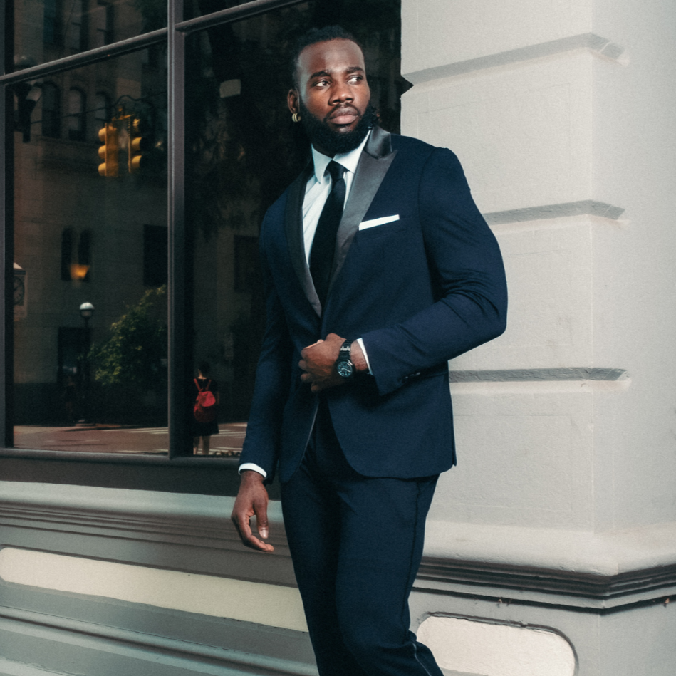 Man in a navy suit standing against a building with a window.