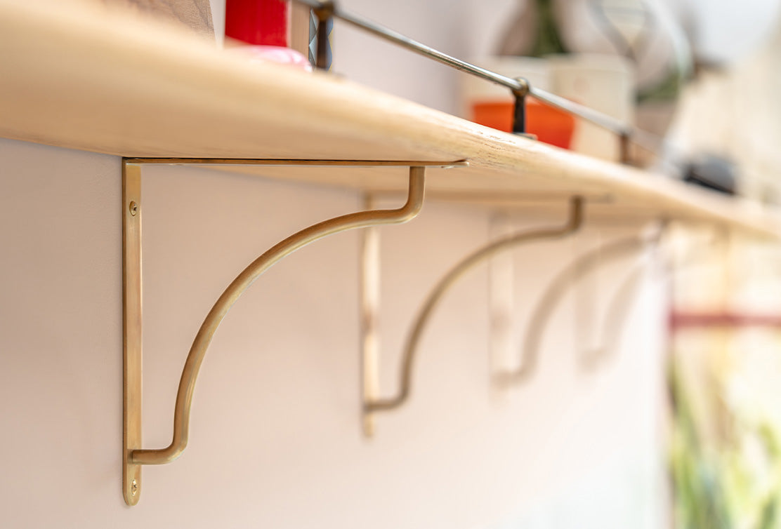 Close-up of antique brass 'Kilburn' shelf brackets supporting a wooden shelf, with blurred background.