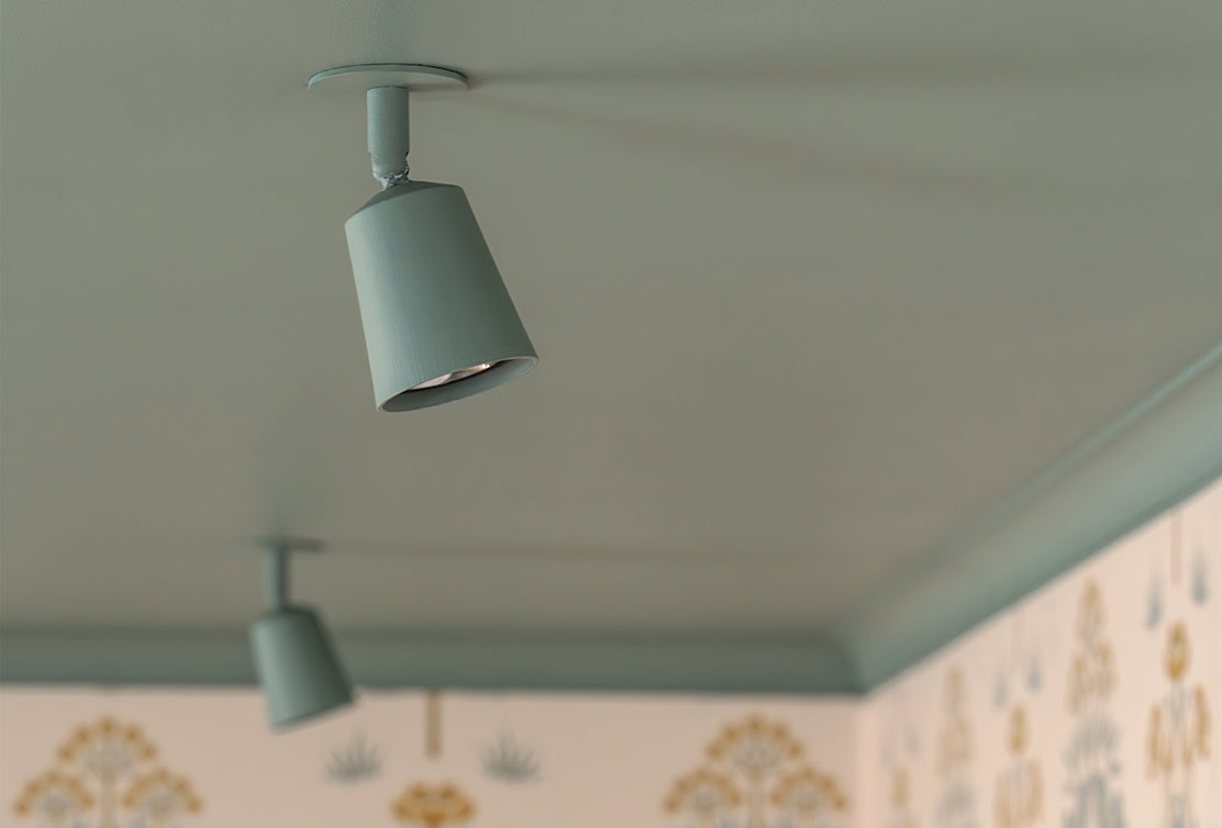 A close-up of elegant, angled ceiling lights on a soft blue-painted ceiling, casting light onto a decorative wallpaper below.