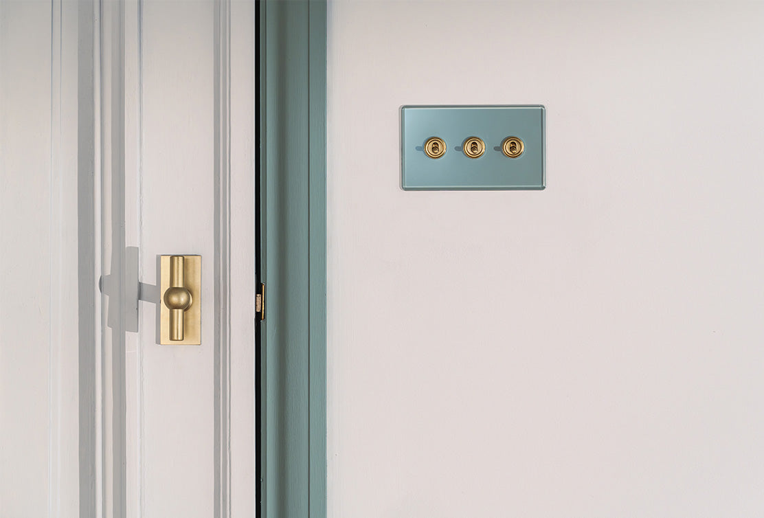 A stylish door corner with a brass knob and a three-button light switch against a pale wall and soft blue trim.