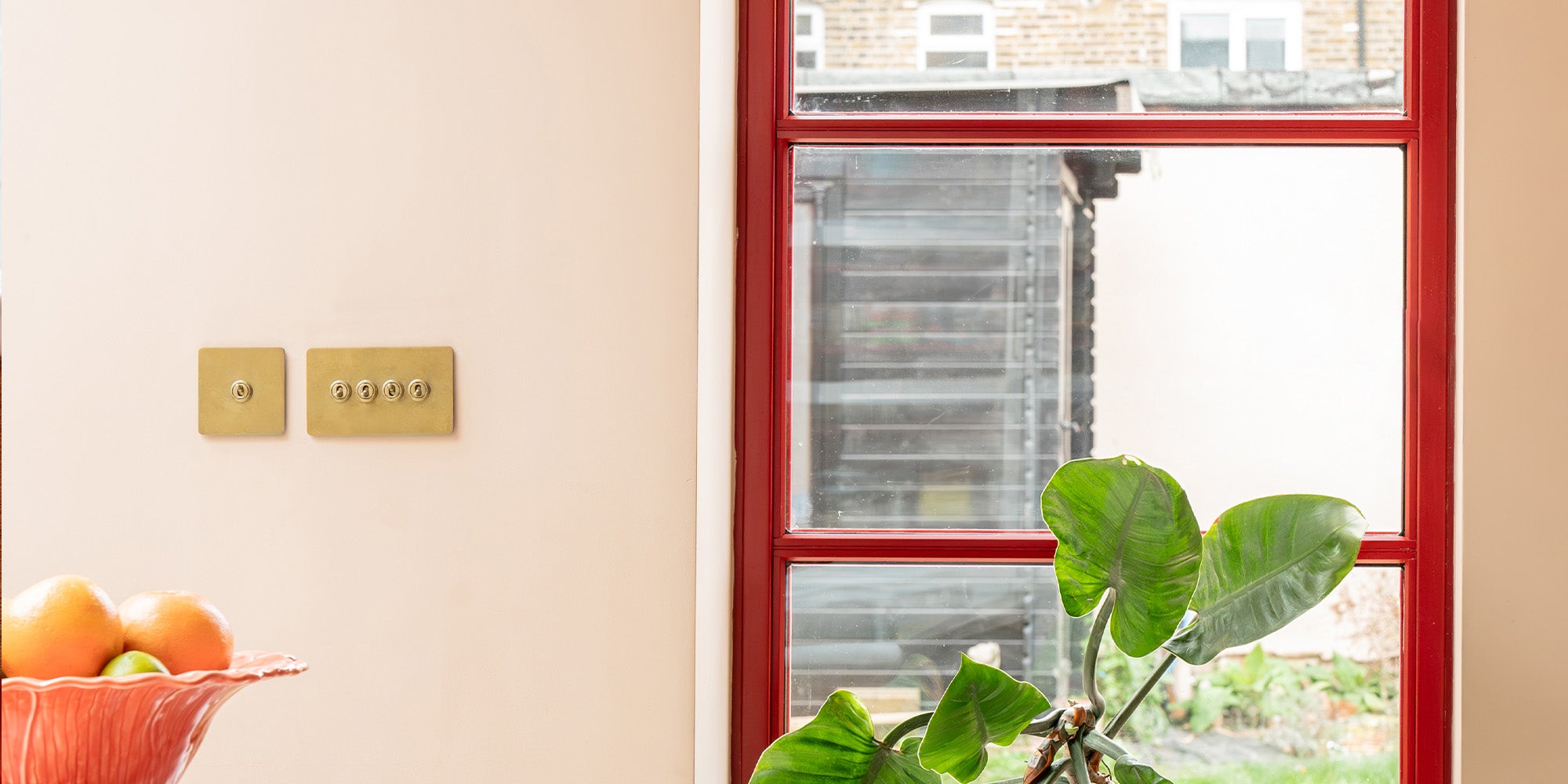 Interior with a red window frame, a pair of antique brass 'Two Way Toggle' switches, and a plant beside a bowl of fruit.