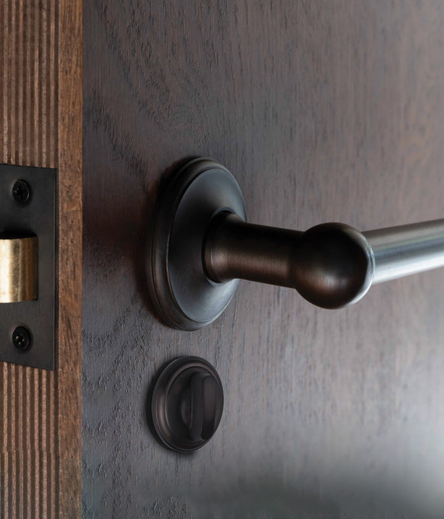 A hand turn a sleek black door handle on a wooden door, with a keyhole visible beside it, set against a blurred outdoor background.