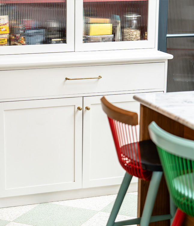 Modern kitchen cabinet with brass handles and colorful chairs in the foreground.