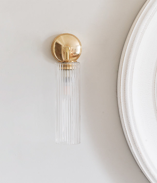 A sleek, brass wall sconce with a ribbed glass shade illuminating a neutral wall beside a round mirror.