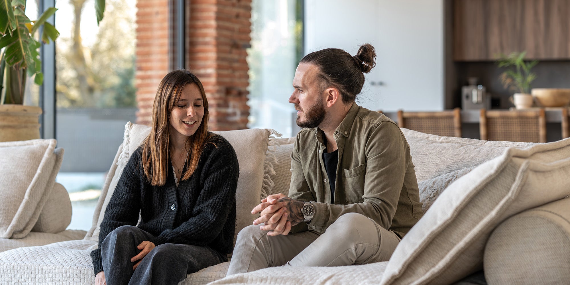 Two people sit on a neutral-toned sofa in a stylish, modern living room with large windows and a relaxed atmosphere.