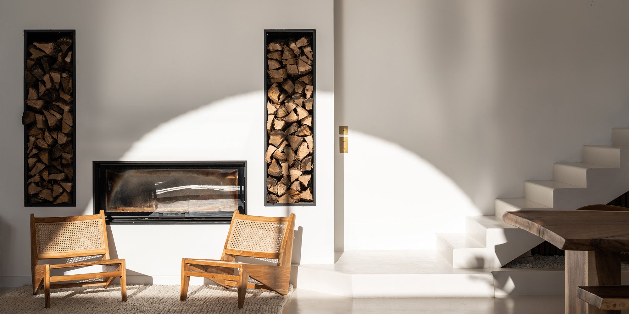 A minimalist living space featuring wood accents, two wooden chairs, a fireplace, and stacked firewood, illuminated by soft sunlight.