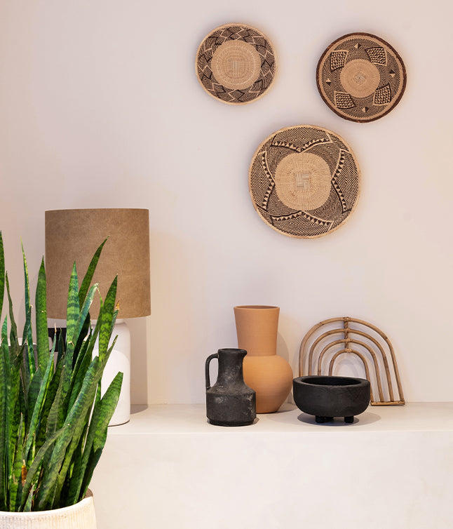 A stylish display of woven wall decor, vases, a pitcher, and a lamp next to a vibrant green plant, set against a neutral wall.