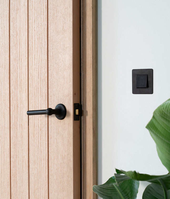 An oak door with a black handle, partially open, next to a black light switch and a green plant in a modern interior.