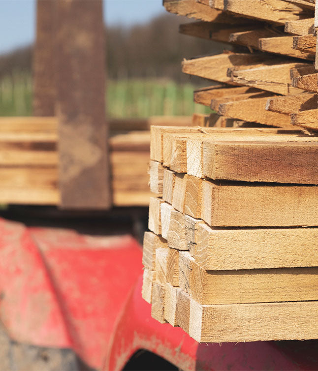 Close-up of stacked wooden planks on a red tractor, with blurred background of green fields and a sunny sky.