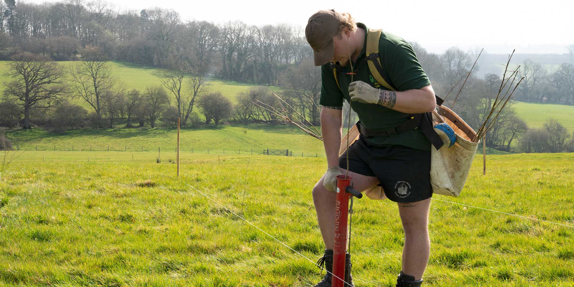 A person in a green shirt and shorts is using a planting tool in a vibrant green field with distant trees and hills in the background.