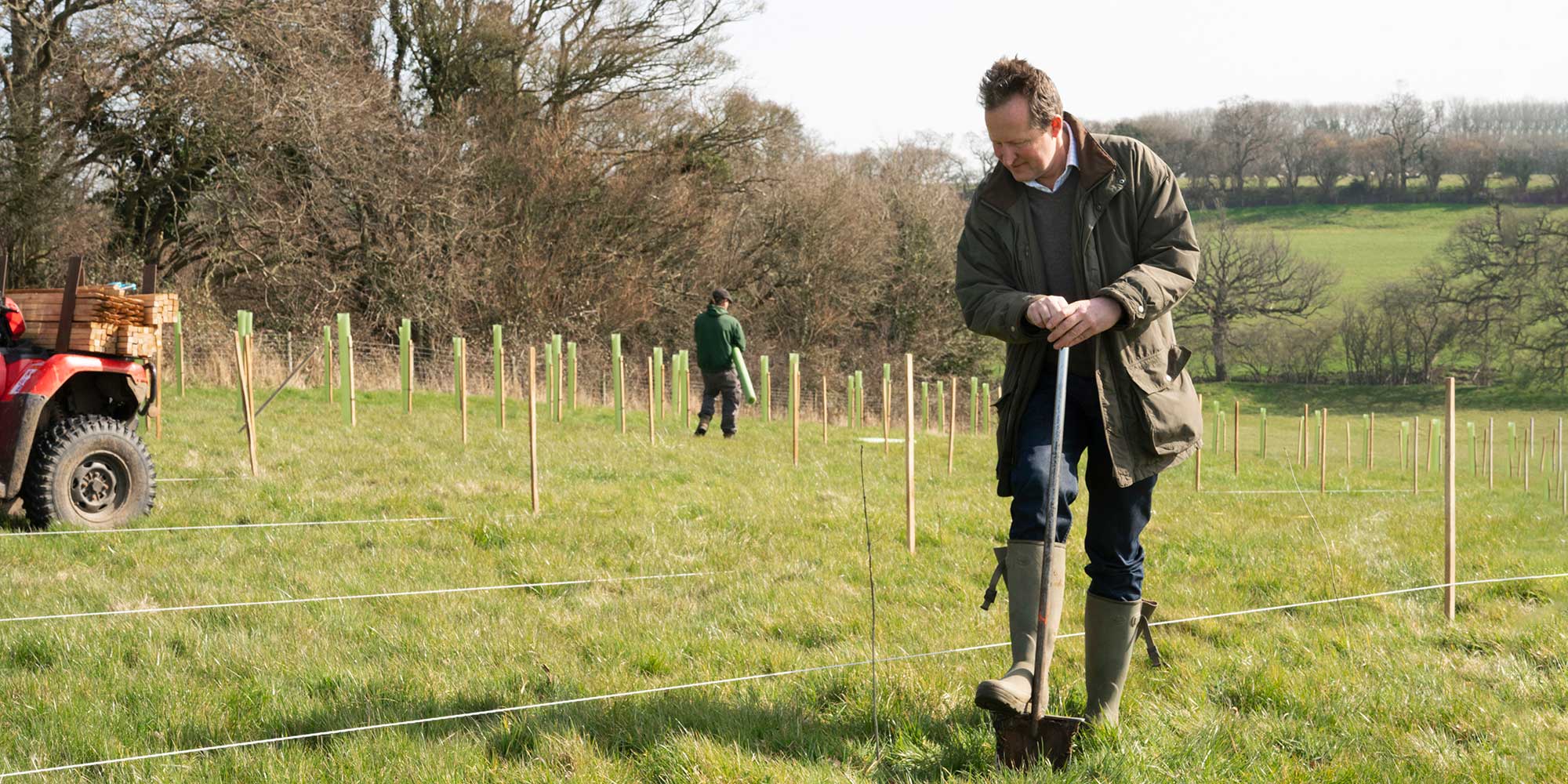 Two individuals work in a grassy field lined with wooden stakes and tubes, with an ATV and trees in the background.