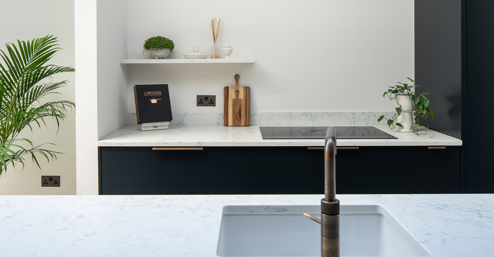 Modern kitchen with marble countertops, a cookbook, wooden cutting board, plant, and sleek black cabinetry. Stylish and minimalist design.
