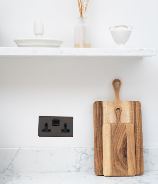 A wooden cutting board stands next to a black electrical outlet, with decorative kitchen items on a marble countertop shelf above.