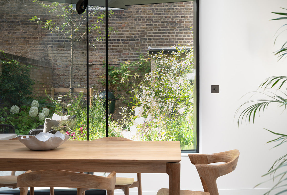 A wooden dining table with a decorative bowl sits indoors, overlooking a lush garden through large glass windows.