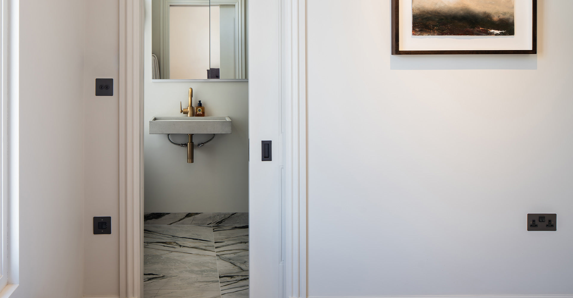 A modern bathroom entrance showcasing a sleek sink with a brass faucet, framed mirror, and stylish marble floor tiles.